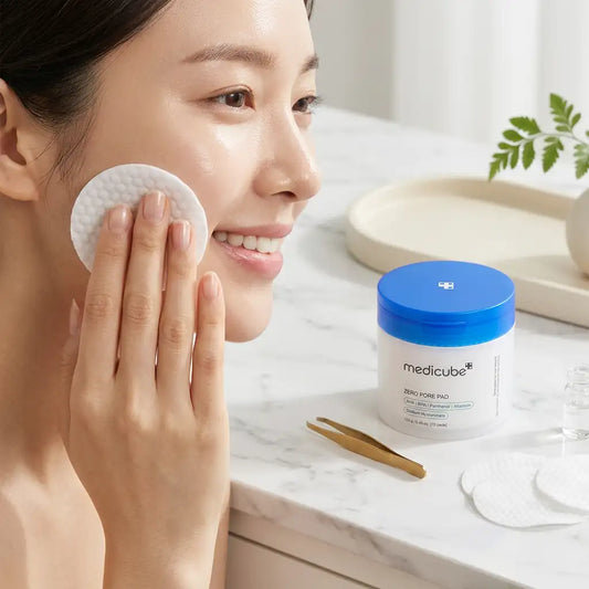 A woman applies a round cotton pad to her face beside a white jar of medicube Zero-Pore Pad skincare product on a marble countertop.