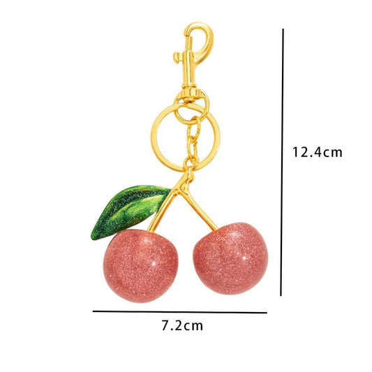 Cherry-shaped keychain or bag charm with two pink glittery cherries, green leaves, and a gold clasp, shown with dimension lines indicating 12.4 cm height and 7.