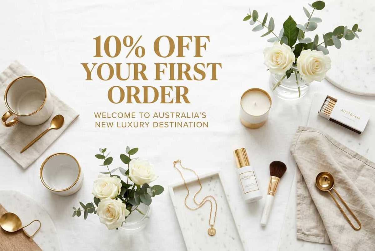 Promotional flat-lay with large text reading '10% OFF YOUR FIRST ORDER' over a styled arrangement of white and gold beauty/coffee props on a light marble surface. Visible items include ceramic mugs with gold trim, spoons, a candle, flowers, a necklace, a brush, and a matchbox labeled Australia.
