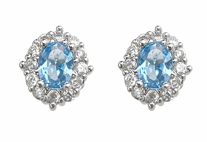 A pair of oval stud earrings featuring light blue gemstone centers surrounded by clear halo stones in a silver-toned setting, shown on a plain white background.