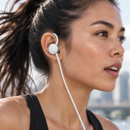 A close-up side profile of a woman wearing a white in-ear earbud with a white cord, outdoors with a blurred cityscape background.