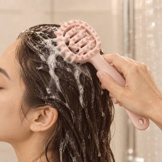A close-up lifestyle shot of a person washing their hair with soapy foam under a shower stream, using a light pink silicone scalp massage brush held by a hand.