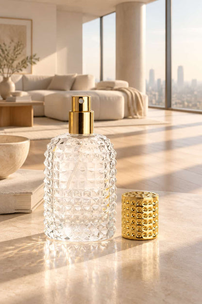 A clear textured glass bottle with a gold spray top sits on a polished stone surface beside a matching small gold textured container in a bright modern loft interior.