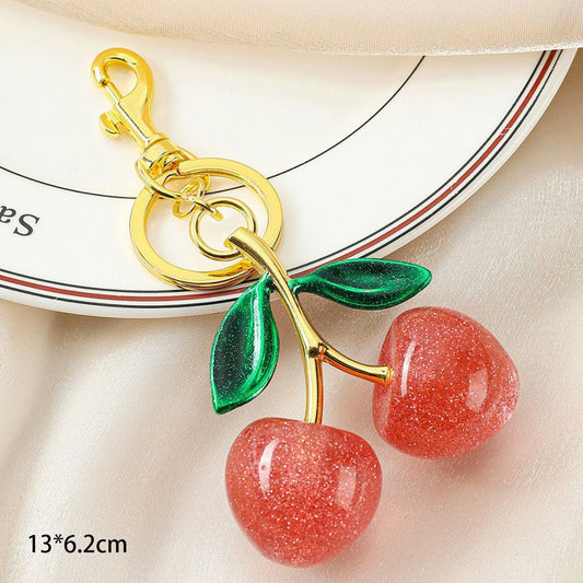 A cherry-shaped keychain or bag charm with two glossy pink-red glittery cherries, green leaves, and a gold clasp, shown on a light fabric surface with a plate edge in the background. Text at bottom left reads 13*6.