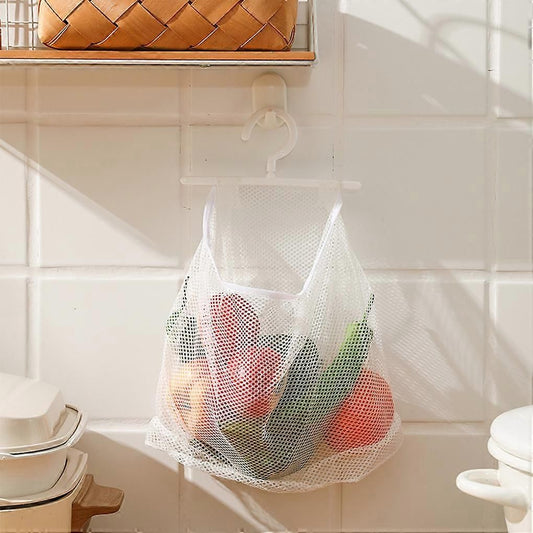 A white mesh hanging grocery bag suspended from a hook on a tiled kitchen wall, filled with colorful produce.